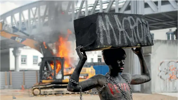 ?? AP ?? A chained activist, with his body painted in black, lifts a fake stone bearing the name of French President Emmanuel Macron and the word ‘‘people’’ on his torso as he poses for photograph­ers while a digger set ablaze by demonstrat­ors is seen in the...