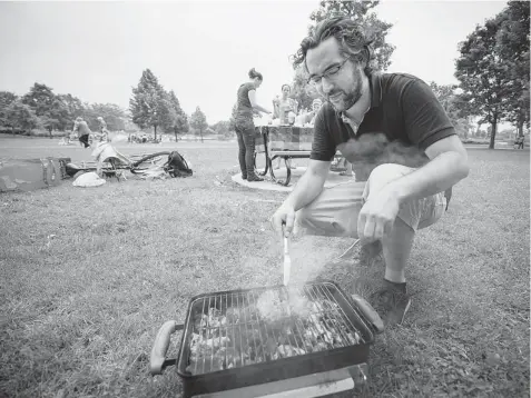  ?? PETER MCCABE/ THE GAZETTE ?? Baptiste Mounie grills up a burger before joining his friends, in background, for a meal in Jarry Park, which has become a haven for barbecue enthusiast­s.