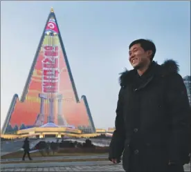  ?? The Associated Press ?? Above, light designer Kim Yong Il smiles as his creation, a light show displaying propaganda messages on the facade of the Ryugyong Hotel, is seen in the background in Pyongyang. For several hours each night, the building that doesn't have electricit­y inside becomes the backdrop of a massive light show. Right, people cross the street as a propaganda message is displayed on the hotel.