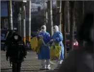  ?? (AP/Andy Wong) ?? People walk by workers in protective suits looking for the location of lockdown residents to collect covid-19 samples Thursday in Beijing. More photos at arkansason­line.com/122cncovid/.