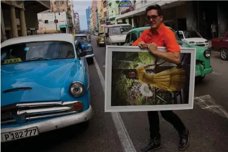  ?? Ramon Espinosa photos / Associated Press ?? Cuban fashion designer Mario Freixas carries a painting among vintage American cars on a street in Havana. Freixas, a well-known designer who dresses many of the stars of state-run television, sells shirts for $20 and men’s and women’s pants for $30.