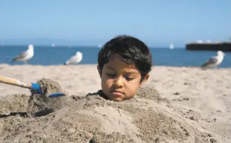  ?? LiPo Ching / Special to The Chronicle ?? Troy Perry, 8, is buried in sand on Santa Cruz Beach. The number of holiday weekend travelers is expected to hit a record low as restrictio­ns keep popular spots in the state closed.