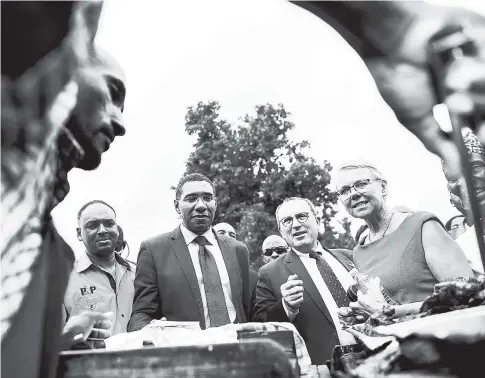  ?? AP ?? In this February 2, 2018 Gleaner photo, jerk chicken vendor Orville Goldson prepares a serving of jerked chicken in the foreground, forming a frame for officials in the background, from left, JSIF Managing Director Omar Sweeny, Prime Minister Andrew...