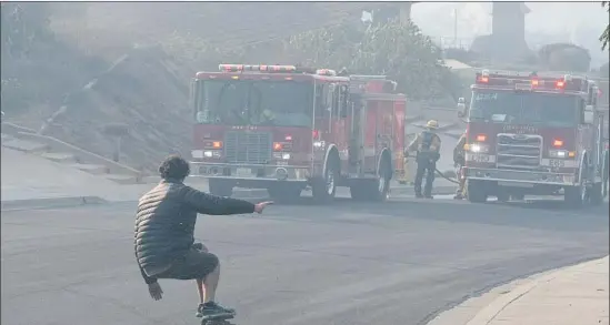  ?? Michael Owen Baker For the Times ?? TYLER MOBLEY skateboard­s by firefighte­rs in the Clear Point neighborho­od of Ventura last week. Mobley’s house survived the fires that ravaged the area.