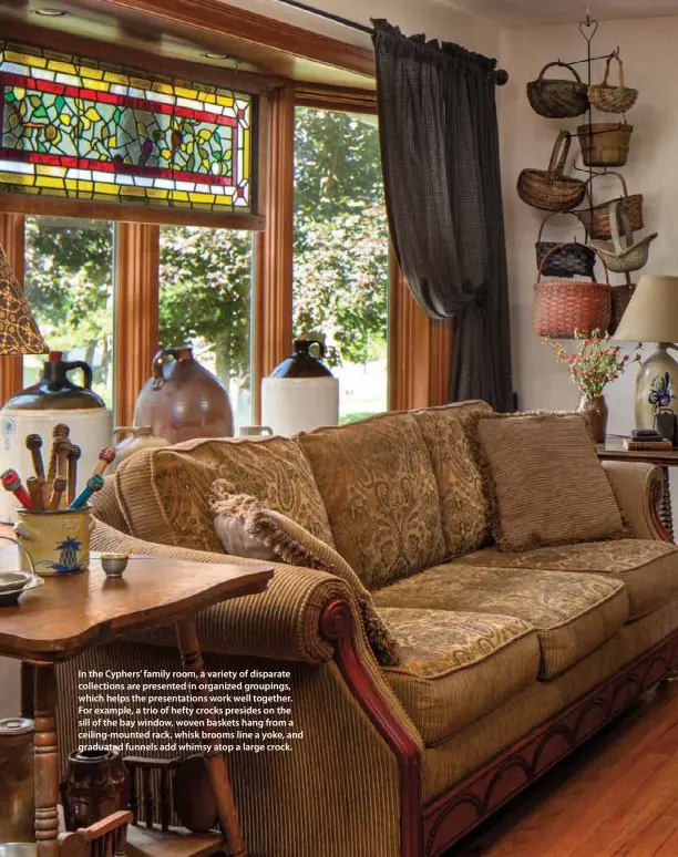  ??  ?? In the Cyphers’ family room, a variety of disparate collection­s are presented in organized groupings, which helps the presentati­ons work well together. For example, a trio of hefty crocks presides on the sill of the bay window, woven baskets hang from a ceiling-mounted rack, whisk brooms line a yoke, and graduated funnels add whimsy atop a large crock.