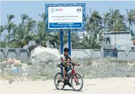  ?? (Ibraheem Abu Mustafa/Reuters) ?? TWO BOYS ride a bike past a USAID sign last week announcing a desalinati­on plant project in the Gaza Strip.