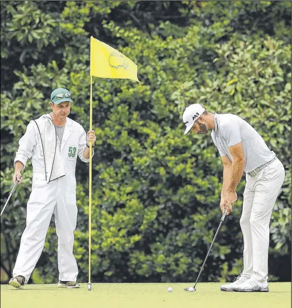  ?? CURTIS COMPTON / CCOMPTON@AJC.COM ?? Dustin Johnson putts on the 11th green while Brooks Koepka’s caddie, Ricky Elliott, tends the flag during their Wednesday morning practice round. Johnson later returned to his rental home, where he fell on the stairs and hurt his back.