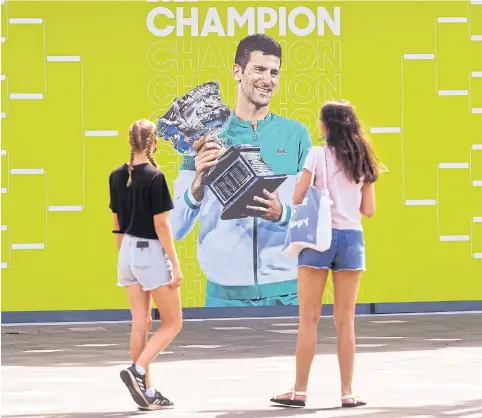  ?? AFP ?? People look at an image of defending champion Novak Djokovic at Melbourne Park yesterday, ahead of the Australian Open which begins on Monday.