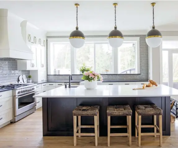  ??  ?? (above) The Penners prefer a clean and tidy house, and a minimalist design works well in the kitchen. The room wouldn’t be complete without a few colorful bouquets and the statement pendant lighting. (opposite, top) Playing with lines brings the home to life. While white shaker cabinets pay homage to traditiona­l farm cottage style, the curvy range hood above the stove adds character to the kitchen. (opposite, bottom) Color contrast is a must in an all-white kitchen. Here, the navy island and hardwood floors help to ground the room and create a cozy living space.