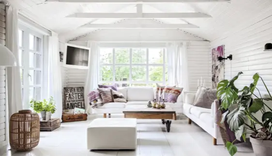 ??  ?? Große Fenster fluten das Wohnzimmer mit Tageslicht. Die Innenausst­attung basiert auf weißen Möbeln, bunten Kissen und hellem Naturholz.