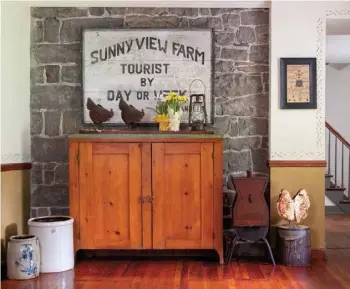  ??  ?? Left: Flanked by a flour sifter and a couple of crocks, the rustic pine cabinet is positioned in front of an original exterior stone wall in the sitting room. The sign, which came with the house, dates from the mid-1900s when the then-homeowner offered shortterm boarding.
Below: The mustard yellow of the kitchen cabinets flows into the adjoining sitting room, which features the same color on the walls below the chair rail and in the hanging quilt and yellowware displayed in the corner cabinet.