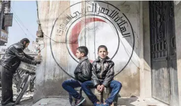  ?? — AFP ?? Syrian children sit before a mural on a wall bearing the logo of the Syrian Red Crescent in the Syrian town of Douma in the rebelheld enclave of Eastern Ghouta on the eastern outskirts of the capital Damascus on Tuesday.