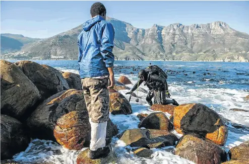  ?? Picture: David Harrison ?? A diver exits the sea along the rocky Hout Bay coastline after poaching perlemoen. A carrier waits to help him with his haul-bag and gear.