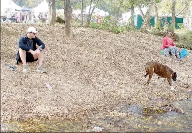  ?? Keith Bryant/The Weekly Vista ?? Brenan Atkinson, left, takes a break from the Spanker Creek Farm Arts and Crafts Festival and watches his boxer, Tyson, check out Spanker Creek. Brenda Pittman, Atkinson’s mother, sits further down the creek’s bank.