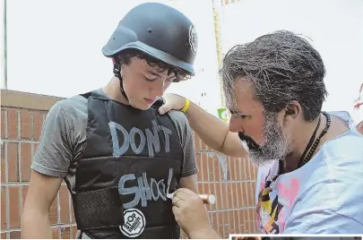  ?? STAFF PHOTOS BY NANCY LANE ?? BACK-TO-SCHOOL FASHION: Parkland shooting victim Joaquin Oliver’s father, Manuel, above right, writes ‘Don’t shoot’ on a protective vest worn by Jack Torres, 16, of Somerville during a fashion show yesterday at City Hall Plaza. Elementary school student Ayden Melendez, 9, right, has his gas mask adjusted by his mother, Nydia Dejesus.