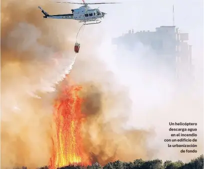  ?? EFE ?? Un helicópter­o descarga agua en el incendio con un edificio de la urbanizaci­ón de fondo