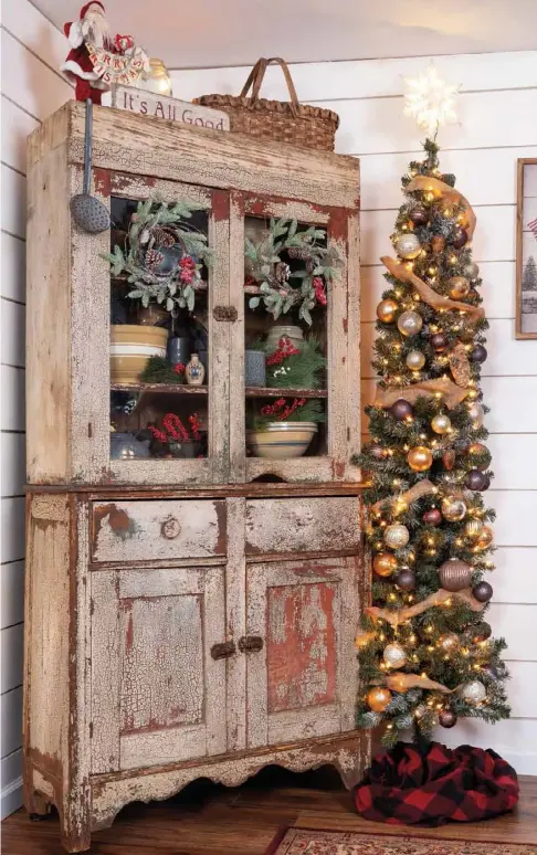  ??  ?? To convert a quiet corner into a charming holiday tableau, Gary angles a tall cupboard into the space and nestles an ultraslim tree to one side. He sprays the tree with artificial snow to match the wreaths on the cupboard doors and then hangs golden ornaments and drapes burlap ribbon, which complement his yellowware and agateware.
For further cohesivene­ss, he interspers­es berry sprays among the wreaths and pottery to pick up the red undertone of the chippy cupboard.