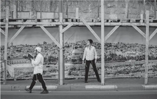  ?? ODED BALILTY/AP ?? Ultra-Orthodox Jewish men walk by a constructi­on site covered with an image of Jerusalem’s old city Friday during a nationwide lockdown to curb the spread of the coronaviru­s.