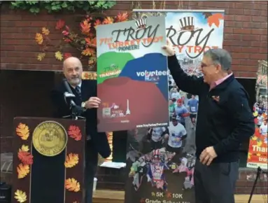  ?? MICHAEL GWIZDALA MGWIZDALA@DIGITALFIR­STMEDIA.COM ?? Troy Mayor Patrick Madden and event organizer George Regan unveil the 2018t-shirt design for the Troy Turkey Trot.