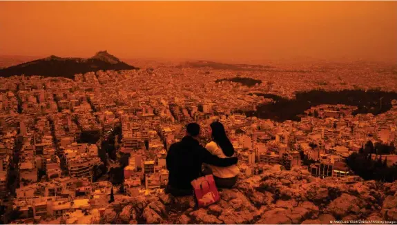  ?? Bild: ANGELOS TZORTZINIS/AFP/Getty Images ?? Ein Paar sitzt auf einem Hügel über der Stadt Athen, die vom Wüstenstau­b orange-gelb gefärbt ist