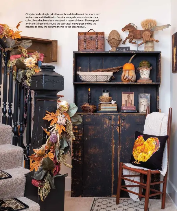  ??  ?? Cindy tucked a simple primitive cupboard sized to suit the space next to the stairs and filled it with favorite vintage books and understate­d collectibl­es that blend seamlessly with seasonal decor. She wrapped a vibrant fall garland around the staircase’s newel post and up the handrail to carry the autumn theme to the second level.