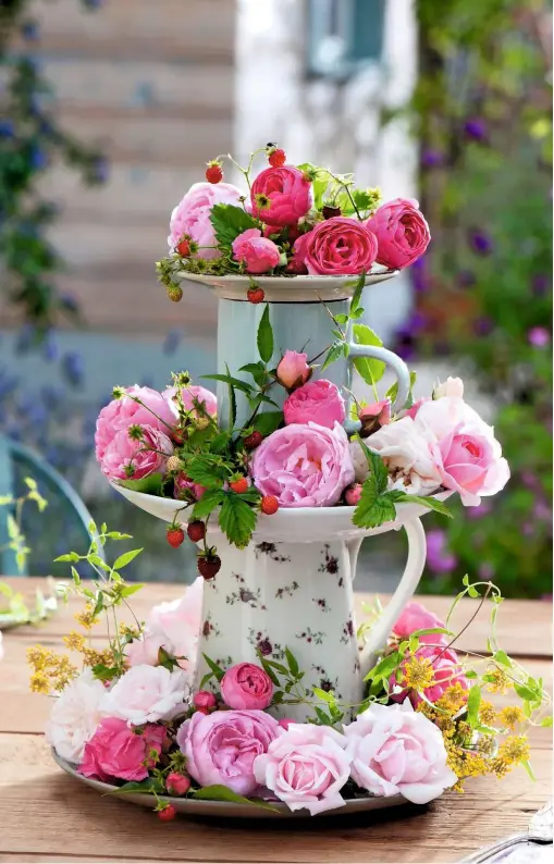  ??  ?? Pretty floral-patterned crockery is piled up to form a tower. Blush pink and crimson roses fill the saucers, their plump blooms interspers­ed with red bead-like fruits.