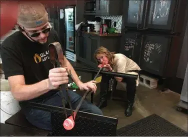  ?? JANET PODOLAK — THE NEWS-HERALD ?? Cathy Knezevich of Mentor puffs air into a blow pipe as Glass Asylum artist Jason Bauck shapes the molten glass with a large cast-iron tweezer.
