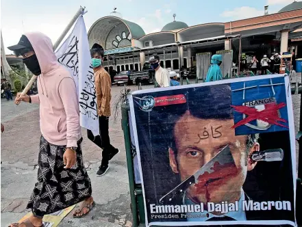  ?? AP ?? Indonesian Muslim men walk past a defaced poster of French President Emmanuel Macron during a protest at Al Jihad mosque in Medan, North Sumatra. Muslims around the world are calling for a boycott of French goods in response to France’s stance on caricature­s of Islam’s Prophet Muhammad.
