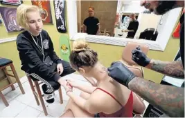  ?? SUSAN STOCKER/STAFF PHOTOGRAPH­ER ?? Justus Culver, 16, a Marjory Stoneman Douglas student, holds the hand of friend Rio Gonzales, 17, a Coral Springs High student, as she gets the number 17 and angel wings tattooed to the back of her neck by artist Ted Mendoza of No Hard Feelings Tattoo...