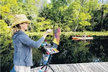  ?? PATRICK CONNOLLY/ORLANDO SENTINEL PHOTOS ?? Artist Manon Sander paints as a kayaker paddles by on the Wekiva River during the Wekiva Paint Out at Wekiva Island.