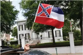  ?? (AP Photo/Rogelio V. Solis) ?? A small Mississipp­i state flag is held by a participan­t during a drive-by “re-open Mississipp­i” protest past the Governor’s Mansion, in the background, on April 25 in Jackson, Miss. This current flag has in the canton portion of the banner the design of the Civil War-era Confederat­e battle flag, that has been the center of a long-simmering debate about its removal or replacemen­t.