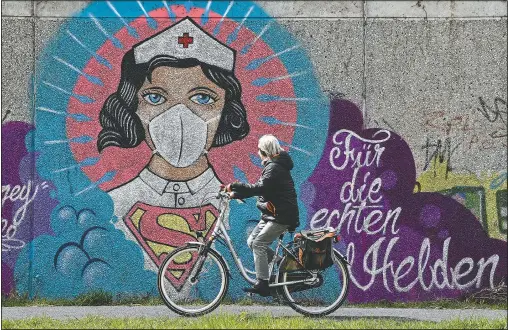  ?? (AP/Martin Meissner) ?? A woman on a bicycle passes coronaviru­s-inspired graffiti by street artist Uzey showing a nurse as Superwoman on Monday in Hamm, Germany. The lettering reads “for the real heroes”.