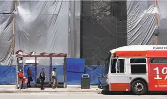  ?? Michael Short / Special to The Chronicle ?? Passengers wait for a Muni bus in front of barriers surroundin­g a constructi­on site on Mission Street and Van Ness Avenue. Bus service has been erratic as major projects are delayed.