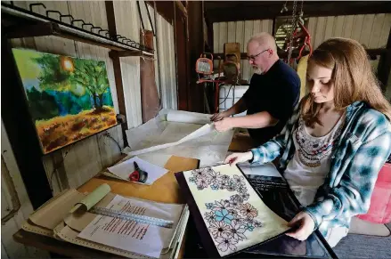  ?? PHOTOS BY CURTIS COMPTON/CCOMPTON@AJC.COM ?? Mart Clamp and his daughter Lauren look over designs, including some of Lauren’s artwork, while consulting at his business on May 8 in Elberton. The painting hanging on the wall of a tree in a forest was one Mart scooped out of the trash after Lauren had thrown it away.