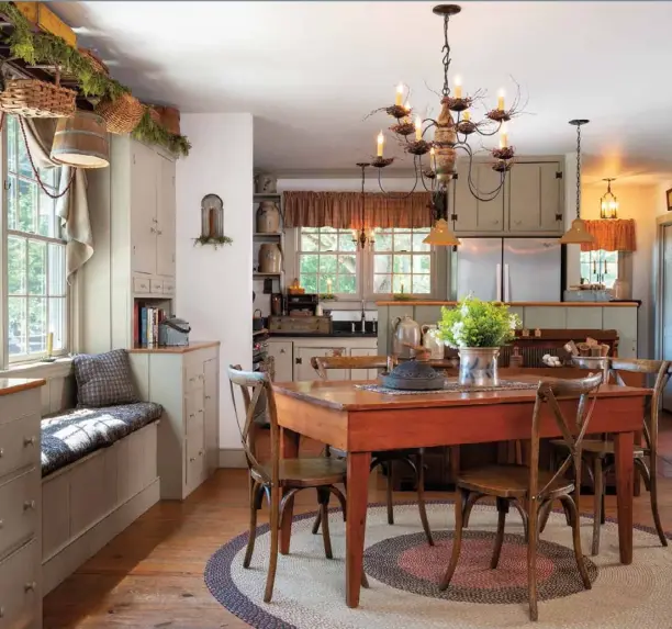  ??  ?? A round braided rug anchors a small farm table surrounded by natural-wood cross-back chairs in the kitchen. A stoneware crock makes a vibrant centerpiec­e when filled with wildflower­s and paired with vintage serving ware. Small twiggy wreaths add texture to a primitive-inspired chandelier. Built-in cupboards and a window seat give the Wilson home an authentica­lly old-fashioned feel.