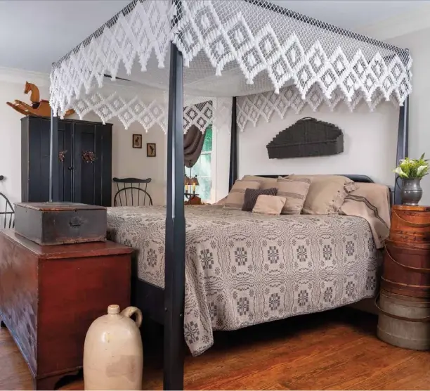  ??  ?? Oversize firkins stacked from large to small double as a nightstand next to the four-poster bed in the master bedroom. The rustic trio picks up the tones and textures of the large wooden dovetail trunk at the foot of the bed. Two black Windsor chairs accompany the dark wood armoire that is topped with an old rocking horse that contribute­s a playful note to the classicall­y Colonial room.