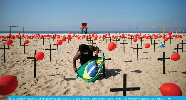  ?? — AFP ?? RIO DE JANEIRO: A demonstrat­or with a Brazilian flag is seen besides crosses during a tribute to COVID-19 victims organized by the Rio de Paz NGO at Copacabana beach on Saturday.