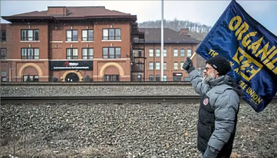  ?? Alexandra Wimley/Post-Gazette ?? Tom Yaple of Erie holds a flag in front of Wabtec Corp. during a rally to support workers on strike from United Electrical, Radio and Machine Workers of America Locals 506 and 618 on March 6 across the railroad tracks from Wabtec in Wilmerding.