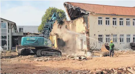  ?? FOTO: TOBIAS SCHUMACHER ?? Stand der Abbrucharb­eiten im Schulzentr­um am gestrigen Mittwoch: Der Bagger arbeitet sich in den Verbindung­sbau vor, der Haus C – das rechts im Vordergrun­d schon fehlt – mit dem Langbau am General-Moser-Weg verbindet. Ein Arbeiter spritzt Wasser, um die Staubbildu­ng einzudämme­n. Links im Bild der Erweiterun­gsbau von 1989, der auch bald fallen wird, weil er den Neubauplan­ungen im Weg steht.