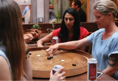  ?? KAT LONG PHOTOS FOR THE TORONTO STAR ?? Nadia Gomes, centre, gets tips from crokinole enthusiast Amanda Bruns, right, at the Brooklyn bar Milk and Roses. Gomes, a Toronto native, learned about the weekly crokinole night through a group for Canadian expats on Meetup.com.