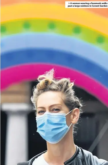  ??  ?? > A woman wearing a facemask walks past a large rainbow in Covent Garden