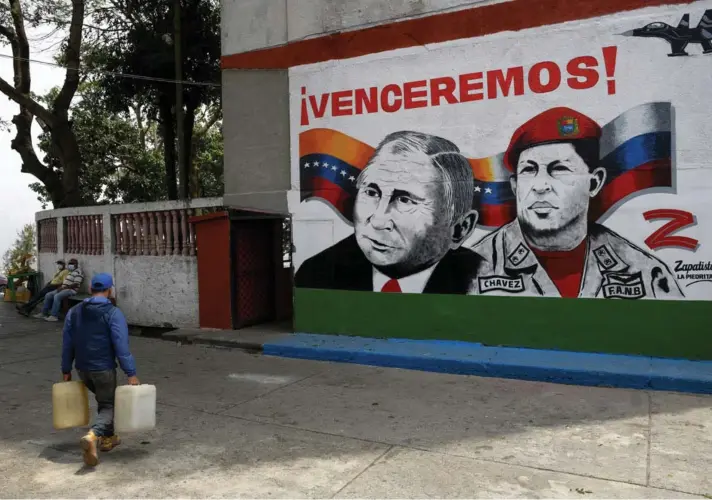  ?? ?? ► Un hombre lleva contenedor­es de plástico frente a un mural que representa al presidente ruso Vladimir Putin y al difunto presidente venezolano Hugo Chávez en Caracas.