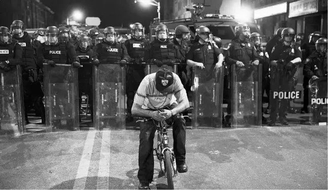  ?? Da vidGoldman/theasociat­e d pres ?? A man sits on a bicycle in front of a line of police officers in riot gear ahead of a 10 p.m. curfew on Tuesday in Baltimore.