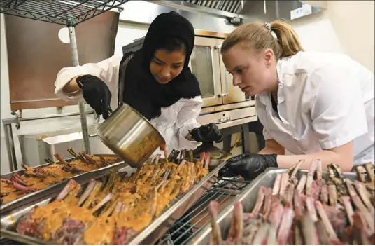  ?? ANDY CROSS — THE DENVER POST ?? Afghan refugee Shukria Mohammad pours sauce over lamb chops with help from University of Denver hospitalit­y management student Jess Bryan in the kitchen at the Joy Burns Center. The Fritz Knoebel School of Hospitalit­y Management program hosted refugees in a five-week food training course called the Ready for American Hospitalit­y program.