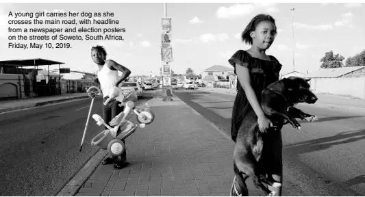  ??  ?? A young girl carries her dog as she crosses the main road, with headline from a newspaper and election posters on the streets of Soweto, South Africa, Friday, May 10, 2019.