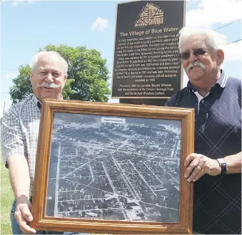  ?? TYLER KULA ?? Gaston Croteau, left, and Serge Gauthier hold an aerial photograph of the former Village of Blue Water in front of a historical plaque marking its spot in southern Sarnia. The former Blue Water residents are in a group organizing a 75th reunion...