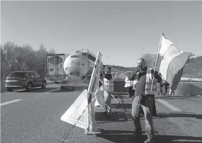  ?? AP Photo/Claude Paris ?? ■ A demonstrat­or wearing a yellow vest waves the French flag while another one sets up a barrier Tuesday on a motorway near Aix-en-Provence, southeaste­rn France. French Prime Minister Edouard Philippe announced a suspension of fuel tax hikes Tuesday, a major U-turn in an effort to appease a protest movement that has radicalize­d and plunged Paris into chaos last weekend.