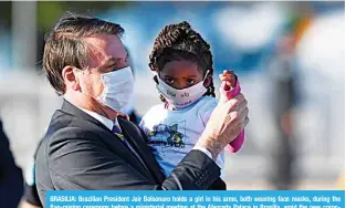 ?? —AFP ?? BRASILIA: Brazilian President Jair Bolsonaro holds a girl in his arms, both wearing face masks, during the flag-raising ceremony before a ministeria­l meeting at the Alvorada Palace in Brasilia, amid the new coronaviru­s pandemic.