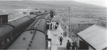  ?? Highland Railway Society Collection/H Davies ?? There is now much activity at the Inverness end of the down platform: passengers alight, barrows are on hand, and unloading has begun. One member of staff is lugging sacks of incoming mail towards the camera, and staff prepare to uncouple the restaurant car from the rear of the train. Meanwhile, having photograph­ed the incoming train passing East box, Roy Hobbs is seen in the field above the downside shelter. The land rising to the left climbs to Fionn Bheinn, a munro of 3,060ft, and the main road to Dingwall goes virtually unseen beyond the goods yard, the railway sharing Strath Bran with the road at this point. The mail will be from Inverness and also from further afield, trains with mail from Glasgow, Perth, and Aberdeen all incoming to Inverness by 10.12am.