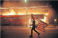  ??  ?? A protester carries a U.S. flag upside, a sign of distress, next to a burning building Thursday, in Minneapoli­s. Protests over the death of George Floyd, a black man who died in police custody Monday, broke out in Minneapoli­s for a third straight night. [JULIO CORTEZ/ THE ASSOCIATED PRESS]